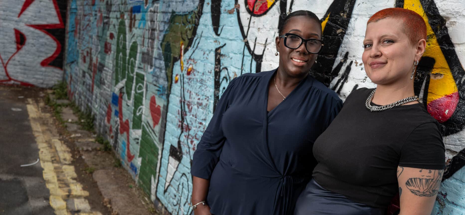 Two women leaning against a graffiti-covered wall