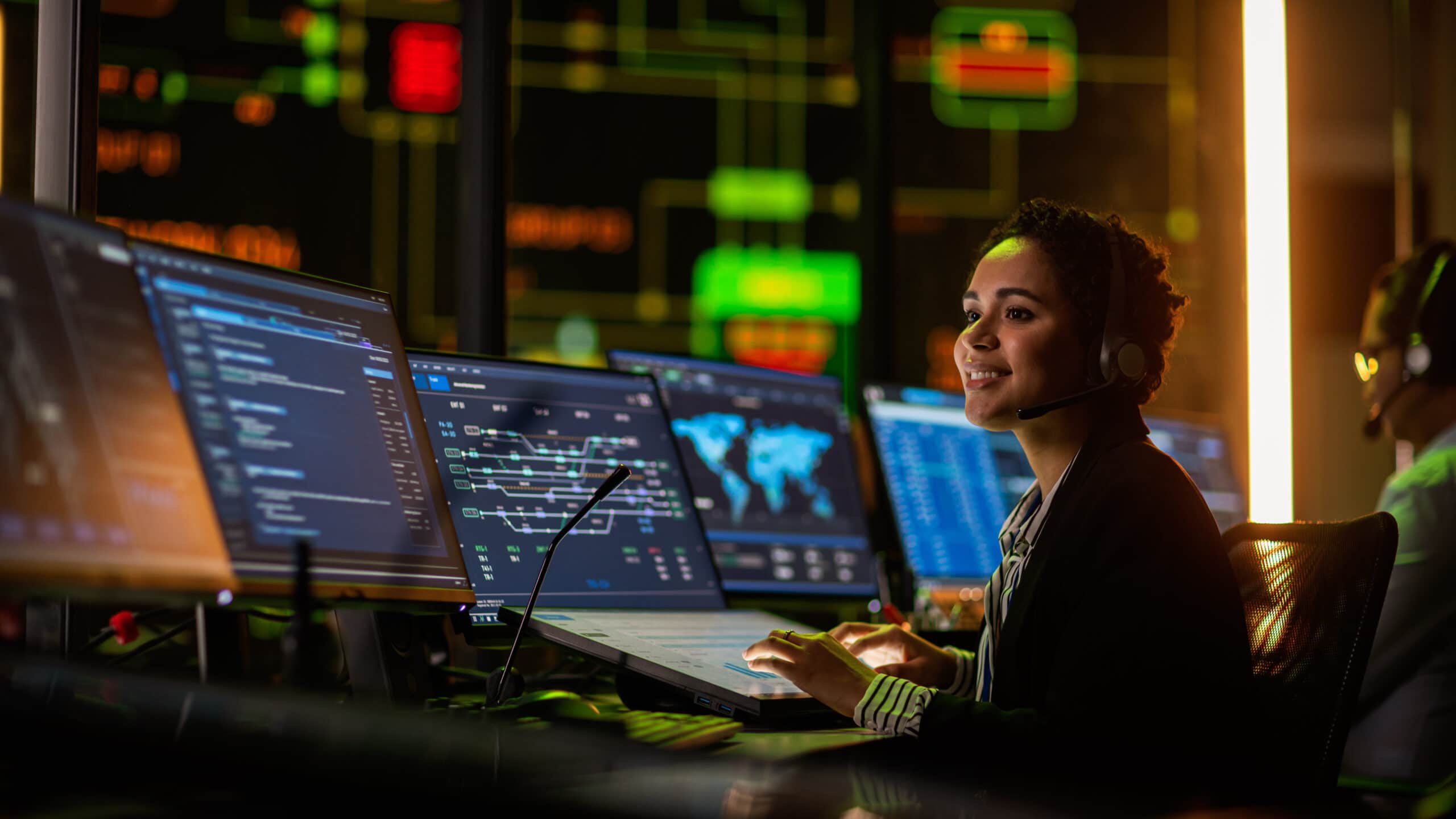 Multiethnic Female IT Technical Support Specialist Talking with a Client in Headphones and Working on Desktop Computer in Monitoring Control Room with Big Digital Screens with Server Blockchain Data.